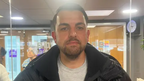 Man with black hair and beard looking at the camera in a black jacket and grey t shirt.