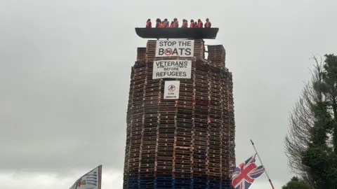 The picture shows a model of a small boat with figures inside on top of a tall bonfire constructed mainly from pallets. Signs saying "Stop The Boats" and "Veterans Before Refugees" are on the bonfire and a Union flag and loyalist flag are at the bottom of the picture