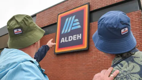 Two men wearing bucket hats, one green and one blue, stand with their back to the camera and point at the Aldi sign, which features the shop's orange and red bordered blue logo and the word Aldeh, on the side of one their brick built store