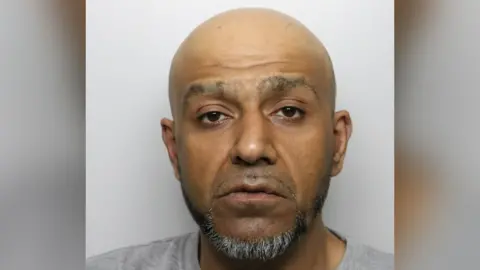 A mugshot of Saqib Khan, with a bald head and a goatee beard.