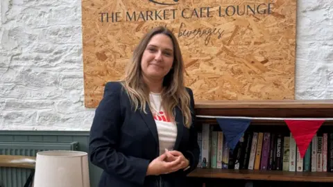 Kate leans against a book case and smiles at the camera. She is wearing a navy jacket and a white t-shirt with red writing on it. She has brown hair with blonde Ombre. Behind her is a wooden sign that reads 'The Market Café Lounge'