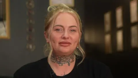 Lucy is sat in a professional piercing studio. Her blonde hair is tied up with two pieces loose, framing her face. She has a floral neck tattoo and piercings on each side of her cheeks and nose. She also has several ear piercings. She is wearing a black t-shirt with a logo on the front. An employee badge is place on the left side of her t-shirt.