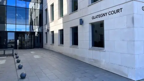 BBC A modern finance building with an angular limestone facade on one wall and glass on another. A sign reads Glategny Court. Small stone spheres line one side of the courtyard.