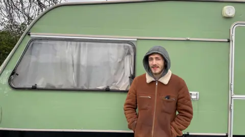 BBC Jack Knight is standing in front of his green caravan, smiling at the camera. He is wearing a brown jacket.