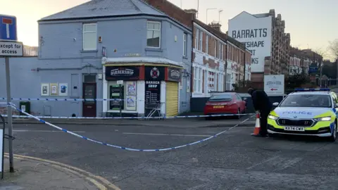 Mark Bulstrode/BBC Police tape blocks off a road. A police car can be seen parked on the outside of the cordon while an officer places a cone on the road.