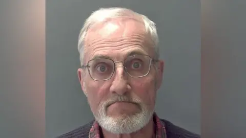Martin Daniels is wearing glasses, and has grey/white hair and a grey/white beard and moustache. He is wearing a red tartan shirt and a ribbed brown jersey.