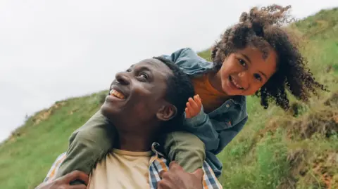 A young girl smiles as she sits on a man's shoulders in the countryside. Both are grinning.