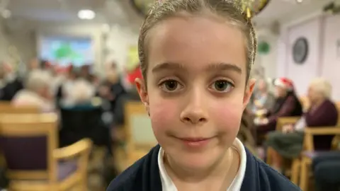 Piers Hopkirk/BBC Head and shoulder shot of a young girl looking to camera in school uniform with elderly people in a room behind her