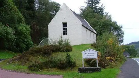 The church is a small, single-storey building with a peaked roof and white walls. There is a long window in the gable end. The church is on a little hill above a road and there is a sign below it that reads St Finnan's.