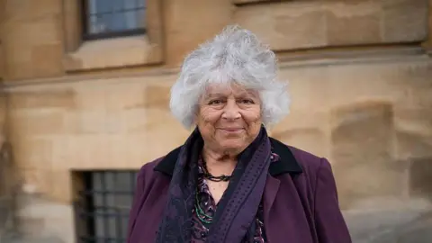 Getty Images Margolyes smiles as she stands in front of a brown building at the Oxford Literary Festival 2025. She wears a purple blazer, a purple blouse, a purple scarf and has short grey curly hair.