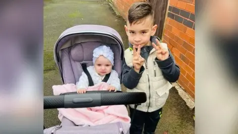 Family handout Sophia is in a pushchair and her brother is standing next to her. She is wearing a white hat with a bow and a white patterned top and is covered by a pink blanket. Her brother is wearing a cream gilet and dark top and trousers. They are on a pavement with a brick wall seen to the right of them.