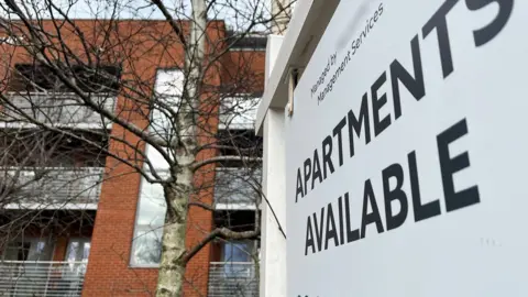 BBC A white estate agent sign hangs from a post. In bold lettering it advertises "Apartments Available". In the background behind a tree there is a red brick, three storey block of flats, with balconies enclosed by glass. Above the building is a sliver of slate grey sky.