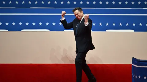 Elon Musk with his fists in the air, wearing a black suit leaving a stage with USA colours in the background.