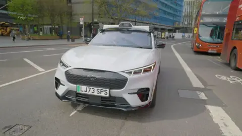 A driverless car on a road in London - there are two red buses to the right