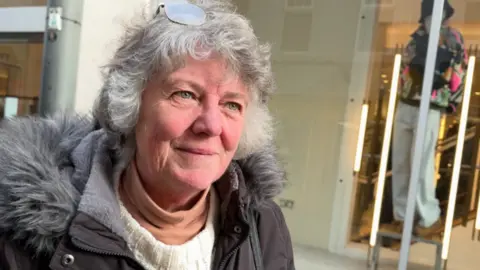 Heather Price has short to medium-length grey hair, with reading glasses resting on her head. She has a parka jacket on with a fluffy trim around the hood and is standing in front of a shop window with a mannequin visible to the right.