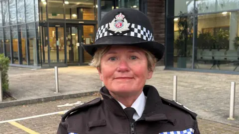 Maggie Blyth stands outside the entrance to Gloucestershire Constabulary's headquarters, which is fronted by very large glass windows. She is smiling slightly as she stands in the car park in her uniform - her body and face are visible from above the shoulders. 