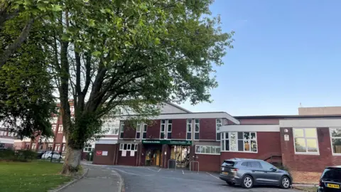 A brick building with dozens of windows. Cars can be seen outside alongside a large green tree.