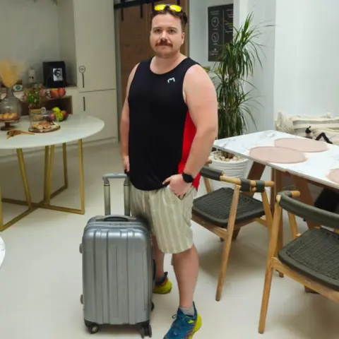 Hywel Roberts Hywel Roberts, wearing vest and shorts and with sunglasses on his head, with a suitcase, in a room