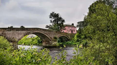 James Johnstone A pink village pub next to a bridge with water flowing underneath it