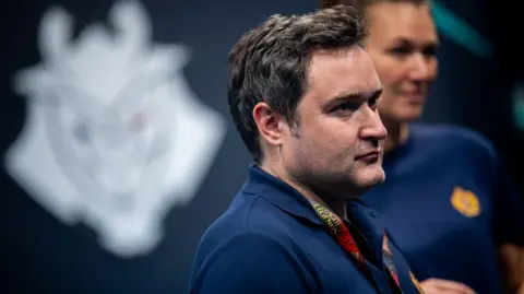 G2 Esports A man with short brown hair looks to the right of the frame wearing a dark blue polo shirt in front of a blurred background.