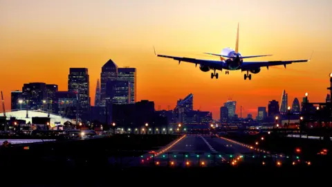 Getty Images: Airplane landing at London City Airport with a row of skyscrapers of the Canary Wharf financial district and the City of London in the background, taken at sunset.