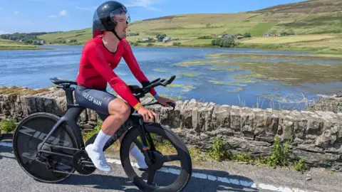 Rob Sorby Tyler Hannay sits on a bike by a lake in front of a green hill. He wears a black helmet, a red top and black shorts. The shorts have Isle of Man written down the side.