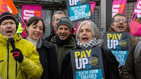 A group of teachers, many holding banners with the words 'Pay Up' in capital letter, are pictured standing outside a red-brick building. 