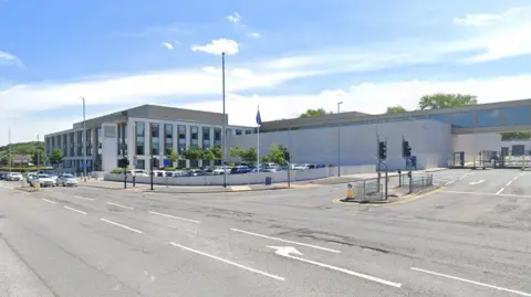 Exterior of Elland Road Custody Suite in Leeds, with the photo taken from the other side of the road. Traffic is being held at traffic lights on a bright, clear day. 