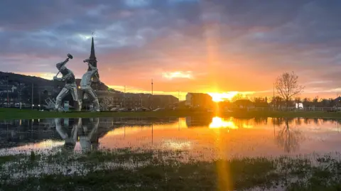 Abigail Duffy A dramatic sunset reflects off a flooded grassy area, with a large metallic outdoor sculpture silhouetted on the left, nearby buildings and a church spire in the distance, and golden light breaking through dark clouds.