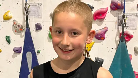 A boy with short blonde hair and wearing a black sleeveless top smiles as he stands in front of an indoor climbing wall.