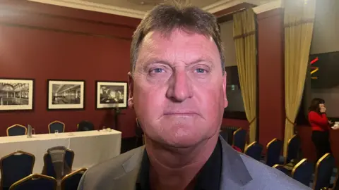 A man with dark hair, wearing a black shirt and a grey jacket