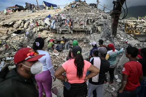 Juan Baretto / AFP via Getty Images A group of people stand with their backs to the camera while search teams navigate the rubble of a destroyed building, in Caraballeda, Venezuela on 22 July.