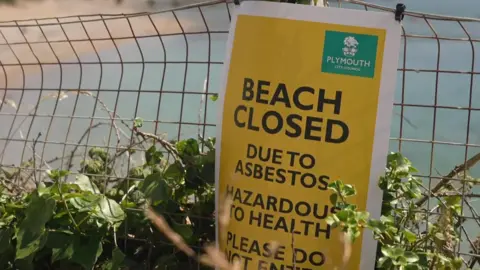An empty Mount Batten beach near Plymouth which has been closed following the discovery of asbestos. There is a yellow warning signing saying "BEACH CLOSED". The sign is attached to a fence and the sea and beach is blurred in the background.