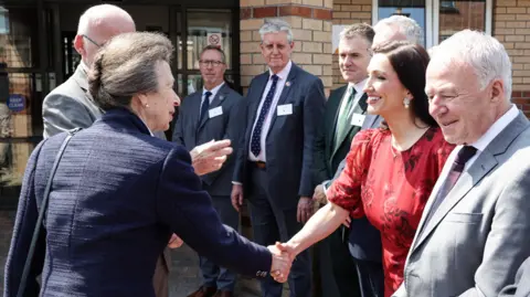 Press Eye The Princess Royal, a woman with grey hair and wearing a blue jacket, greets Emma Little-Pengelly, a woman in a red dress, who has dark hair. There are other men with grey hair in the photo.