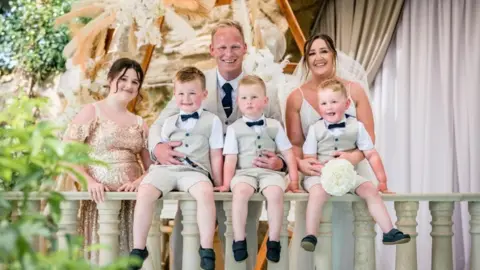 Lauren A posed wedding photo. A man wearing a three piece light coloured suit and dark tie has his arm round two young boys with light hair wearing matching suits and dark bow ties. On the right is a brown haired woman in a white wedding dress and veil, with her arms around a third boy in the same outfit. All three boys are sat on a grey bannister with their legs hanging over the side. On the far left is a teenage girl with brown hair in a gold sparkly bridesmaid dress.