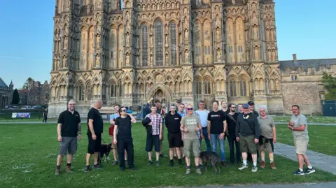 A group of men stand in front of Wells Cathedral smiling. It is a sunny weekend and the ornate facade of the cathedral is behind them.