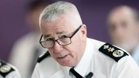 PA Media Jon Boutcher has light grey hair and is wearing black-framed glasses. He is wearing his police shirt and a black tie