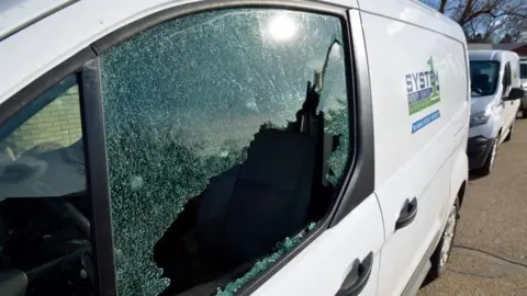 A white van with its window smashed. Another two white vans can be seen parked behind it in a row.