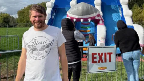 Angus, left, wearing a white T-shirt, standing in front of a large blue inflatable ride with people watching the children using the ride behind him