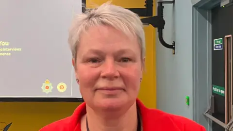 A woman with short blonde hair and a red jacket is smiling as she stands in front of a projector screen.