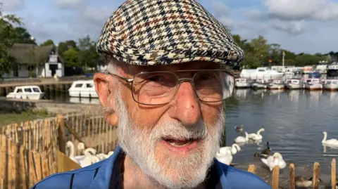 Luke Deal/BBC Laurie Rookie smiles at the camera. He wears a chequered flat cap and blue shirt. He has a grey beard and wears glasses.