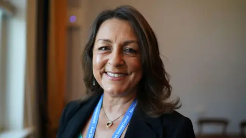 PA Media A woman in a black suit and turquoise top is smiling.