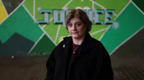 Reuters Kimia Zabihyan, a woman with short brown hair wearing a black fleece and maroon top, stands in front of a green mural that says 'justice'.