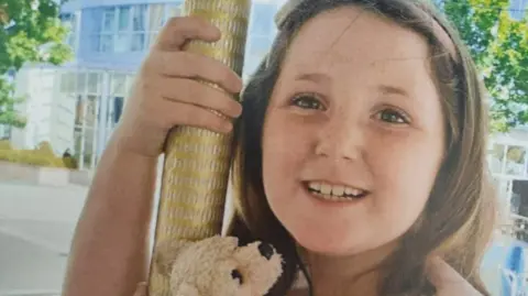 Minnie-Rae Dunn smiles at the camera. She had brown hair held back by a band and carries a small teddy bear.
