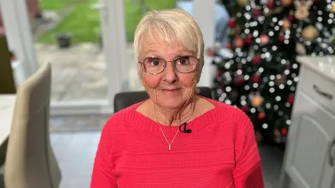 Audrey Hayter sitting in her home with patio doors in the background, looking out onto a lawn. She is wearing a bright pink/red knitted top and wearing glasses.