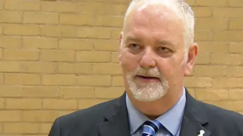 Jonathan Evison, the police and crime commissioner for Humberside has short white hair and a short white beard and moustache. He is looking to the side and wearing a black blazer, a light blue shirt and a blue stripy tie. A yellow brick wall can be seen behind him.