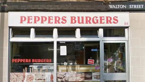 The front of Peppers Burgers. Which has it's name in red on a white sign in front of a large glass facade.