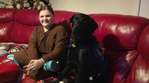 Scarlette, wearing a brown tracksuit, sits in her sitting room on a red sofa with her black Labrador support dog, called Ivanhoe.