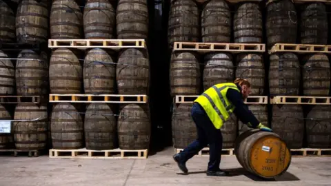 Stacked whisky casks and a man in a high vis jack rolling a cask in the foreground