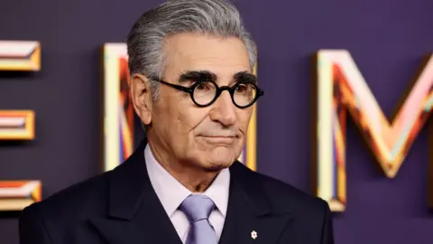 Getty Images Eugene Levy menghadiri Emmy Awards dengan mengenakan dasi ungu, kemeja merah muda, dan setelan gelap